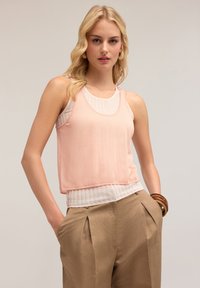 Top fără mâneci, cu un strat superior transparent de culoare roz deschis și un strat interior fin, alb, combinat cu pantaloni plisati maro.