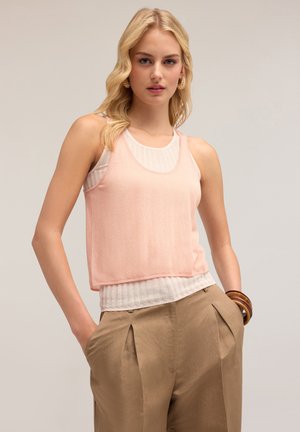 Ärmlös topp med ett ljust rosa, genomskinligt ytterlager och ett ribbat vitt innerlager, kombinerat med bruna plisserade byxor.