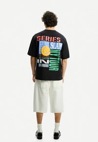 Black oversized t-shirt with colorful, multi-font graphics. Features a tennis ball graphic and text elements. Worn with cream shorts and black shoes.