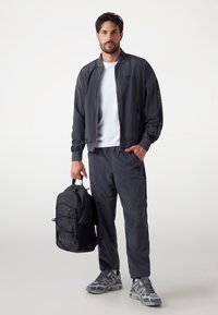 Dark gray bomber jacket with subtle texture, white T-shirt, matching pants, and black backpack. Sneakers feature gray and blue accents.