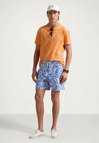 Άντρας με πορτοκαλί μπλουζάκι, μπλε και άσπρα λουλουδάτα μαγιό, άσπρο καπέλο, γυακιά ηλίου κρεμασμένα στον γιακά, άσπρα sneakers και ρολόι στον καρπό, στέκεται.