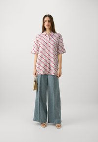 Cămașă cu mânecă scurtă și guler, cu un model geometric roz și roșu, asociată cu pantaloni largi din denim și o geantă mică bej.