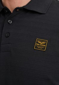 Zwarte polo gemaakt van textuurstof met een drie-knopen sluiting. Voorzien van een gele en zwarte label met "PME LEGEND" en een decoratief design.