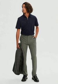 Jūras spēku puszipa apģērbs, zaļi bikses, melni oficiālie apavi un pelēks pārbaudīts žakets, kas tiek turēts. Modelis ar cakotām matiem pozē.