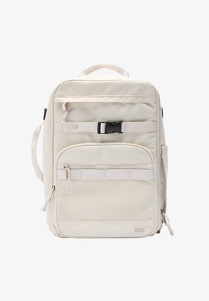 Beżowy plecak wykonany z tkaniny, z górnym uchwytem, regulowanymi szelkami, wieloma zamykanymi kieszeniami oraz przednią sekcją z siatki.
