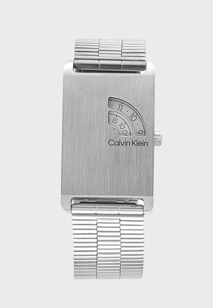Montre-bracelet argentée Calvin Klein avec cadran rectangulaire, petites fenêtres courbes affichant les heures et les minutes, et bracelet à maillons en métal.