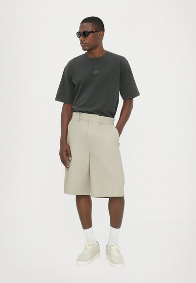 T-shirt gris foncé avec logo, shorts beiges, chaussettes blanches et baskets claires. Mannequin portant des lunettes de soleil noires. Coupe simple et décontractée.