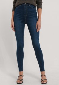 Tmavě modré skinny džíny s vysokým pasem vyrobené z denimu, s hladkou texturou a přiléhavým střihem, doplněné černými sandály na podpatku se špagetovými pásky.