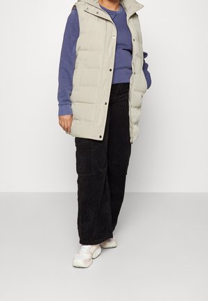 Personne portant un gilet matelassé beige sans manches par-dessus une chemise à manches longues bleue, un pantalon large en velours côtelé noir et des baskets blanches, debout.
