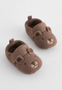 Papuci pentru bebeluși, împletiți în nuanțe de maro, cu un design al feței de urs. Dispun de o curea cu velcro, vârful rotunjit și un material texturat pentru aderență.