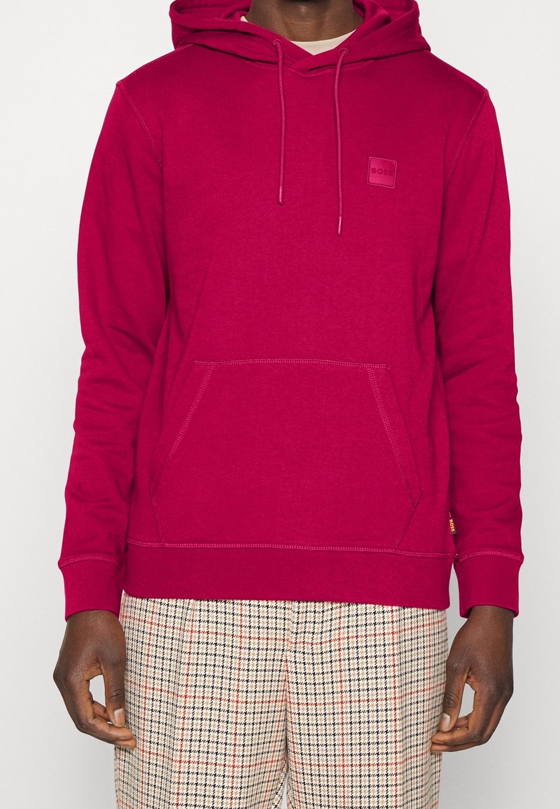 Sweat à capuche rouge avec une poche kangourou et une capuche à cordon de serrage ; comporte un petit patch logo en relief. Associé à un pantalon à carreaux.