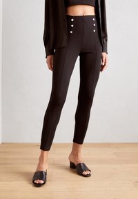 Leggings negros de talle alto con textura suave, con tres botones decorativos en la cintura. Llevados con sandalias negras deslizantes.