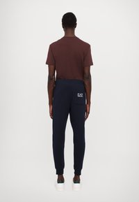 Sötétbarna póló, amelyet tengerészkék melegítőnadrággal kombináltak, melynek hátsó zsebe és logó részlete van, puha pamutkeverékből készült. Fehér lábbeli látható.