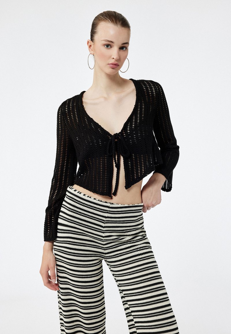 Schwarze, gestrickte, verkürzte Strickjacke mit langen Ärmeln und einer Vorderknopf-Verschluss, kombiniert mit schwarz-weißen, gestreiften, weiten Hosen.