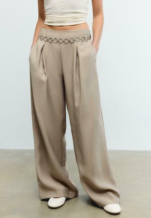 Beżowe spodnie o szerokich nogawkach z plisami i paskiem z łańcuchów, zestawione z białymi butami i jasnym, krótkim topem na jednolitym tle.