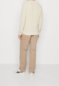 Krämfärgad stickad tröja med ett texturerat mönster, kombinerad med beige byxor och vita sneakers med svarta och grå accenter.