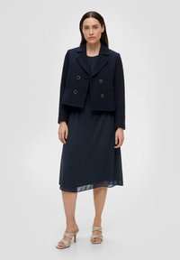 Navyblauwe textuur cropped jack met dubbele rij knopen, gecombineerd met een vloerlange navy jurk. Model draagt lichte sandalen met bandjes.