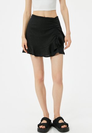 Jupe mini noire à volants avec un design froncé sur le côté, fabriquée à partir d'un tissu texturé. Modèle portant des sandales noires à lanières.