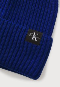 Kék kötött sapka bordázott felülettel, fekete címkével, rajta a "Calvin Klein Jeans" felirattal és egy stilizált "CK" logóval.
