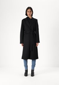 Svart ullblanding lang frakk med spiss krage, belte i midjen og fulle ermer, kombinert med lyseblå jeans og svarte ankelstøvler.