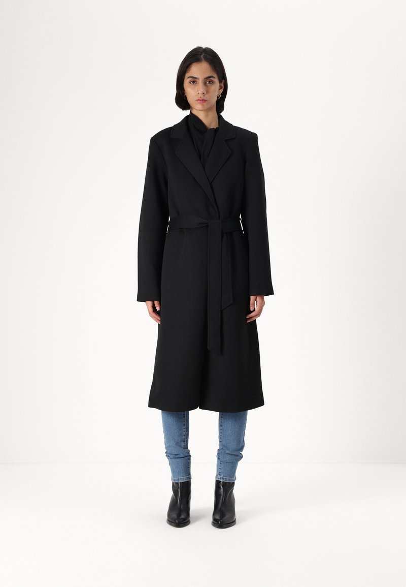 Svart ullblanding lang frakk med spiss krage, belte i midjen og fulle ermer, kombinert med lyseblå jeans og svarte ankelstøvler.