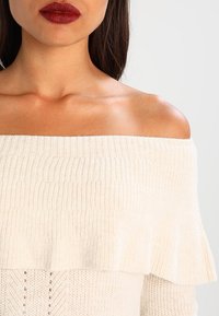 Cremfarbener Pullover mit schulterfreier Form, geripptem Textur und geschichteten Volants. Mit lockerer Passform und weichem Stoff.