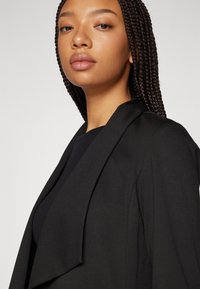 Svart blazer med en strukturert revers, laget av glatt stoff. Har en skreddersydd passform, minimal detaljering og en ren silhuett.