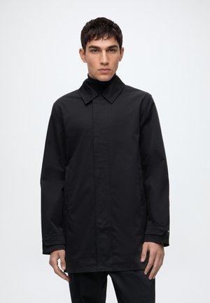 Homme portant un manteau noir à manches longues avec un col, debout devant un fond clair uni, regardant vers l'avant.