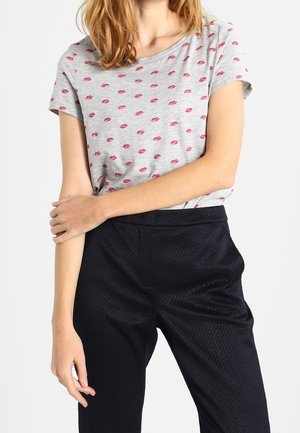 Kvinde iført en grå T-shirt med korte ærmer med røde læbestifttryk og teksturerede sorte bukser med høj talje, armene krydset om taljen.