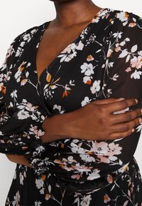 Robe noire à fleurs avec un tissu texturé et fluide. Présente des fleurs blanches et orange, un décolleté en V profond, et des manches bouffantes avec des poignets froncés.