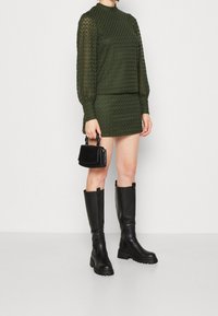 Suéter de punto texturizado verde con mangas largas y cuello alto, combinado con una mini falda texturizada a juego. Bolso negro y botines hasta la rodilla.