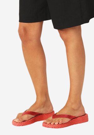 Gambe con pantaloncini neri e infradito rossi con suola piattaforma testurizzata e cinturini larghi, su sfondo bianco.