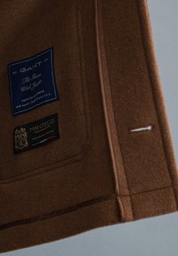 Braune Wolljacke mit einem blau-schwarzen Etikett, das Marke und Pflegehinweise zeigt. Mit sichtbaren Nähten und einem seitlichen Knopfloch.