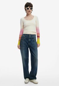 Enganliegendes cremefarbenes Oberteil mit Ärmeln in einem pink, orange und gelb verlaufenden Muster; lockere blaue Jeans; weiße Sonnenbrille; weiße Schuhe.