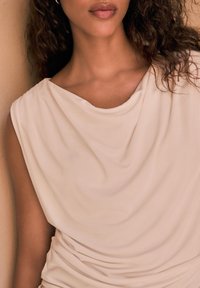 Top beige drappeggiato con una texture morbida e liscia. Presenta un collo a cisterna e dettagli arricciati in vita, che accentuano la silhouette.