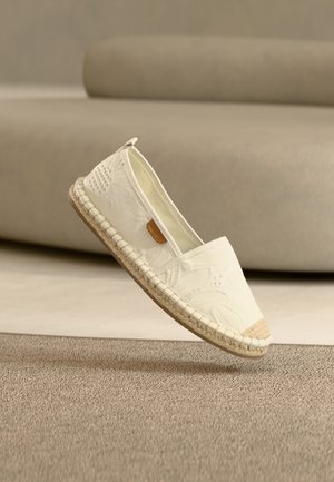 Biała espadrylka z teksturowanej tkaniny, z przeszywaną jutową podeszwą i małą brązową metką, ustawiona na podeszwie na beżowym tle.