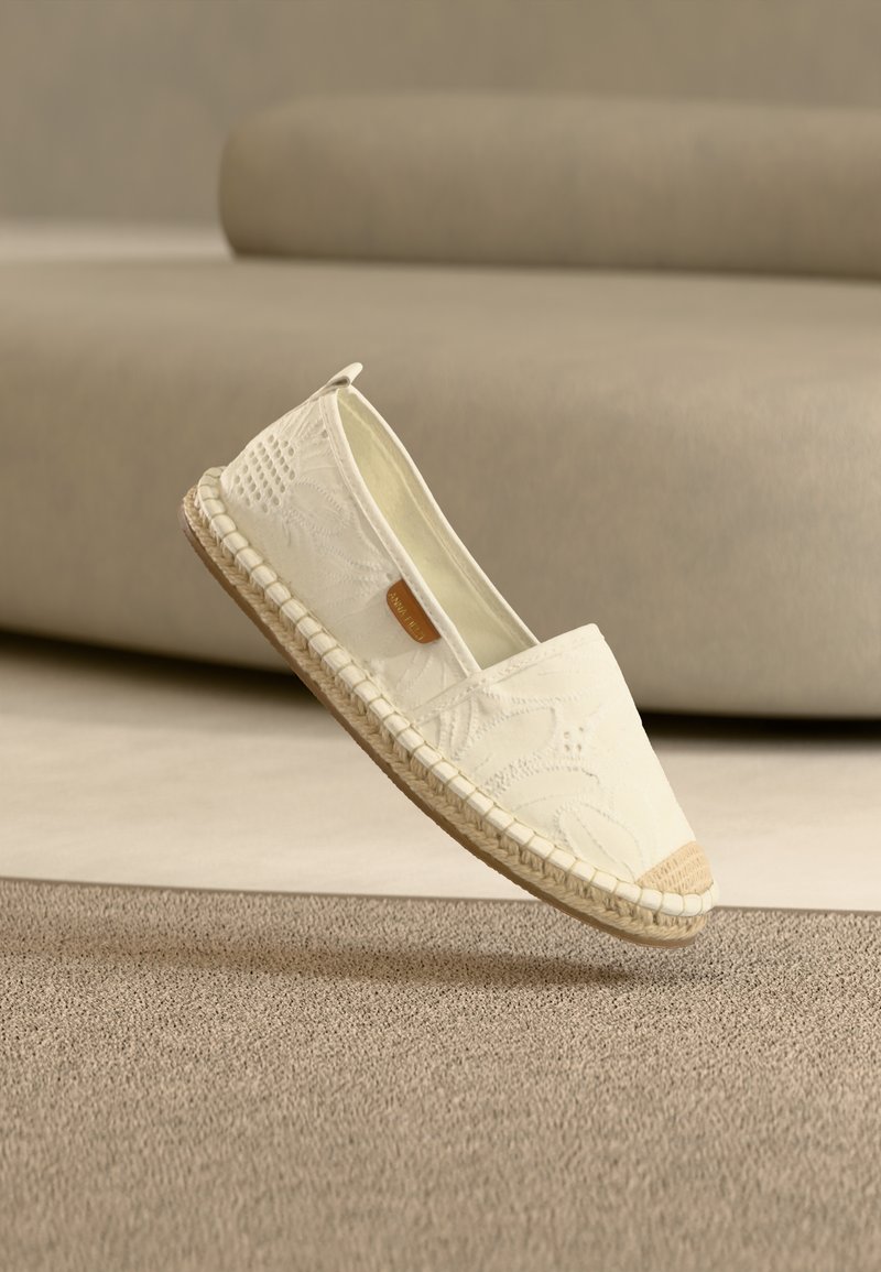 Λευκό παπούτσι εσπαντρίγια με υφασμάτινη υφή, ραμμένη σόλα από τζούτ και μια μικρή καφέ ετικέτα, ισορροπημένο στη σόλα του πάνω σε μπεζ φόντο.