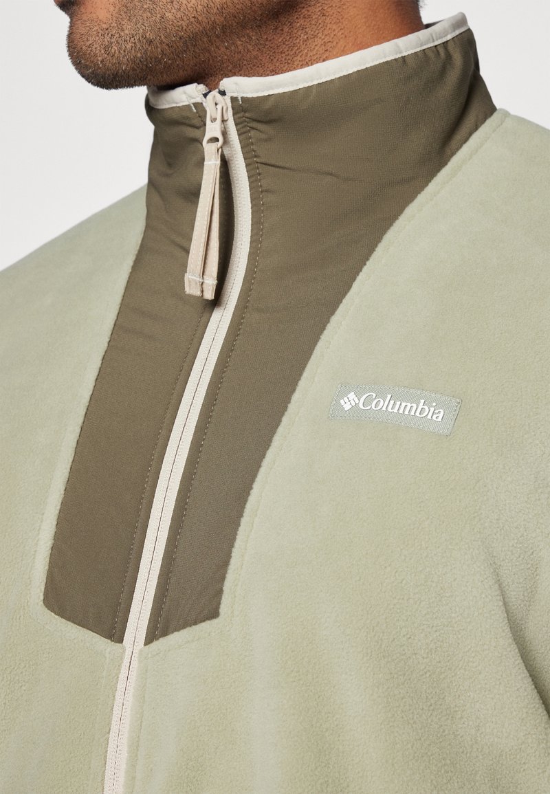 Veste en polaire de couleur vert clair avec un panneau vert foncé. Elle possède un col montant, une fermeture éclair à l'avant et un logo Columbia sur la poitrine.