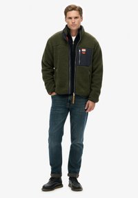 Giacca in pile verde con tasca sul petto blu navy, chiusura con cerniera e tasche laterali, indossata sopra una maglia nera e abbinata a jeans scuri e stivali.