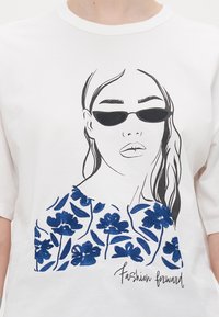 Hvit t-skjorte med en svart linjetegning av en kvinne med solbriller og blå broderte blomster, med teksten "Fashion forward" under.