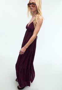 Robe maxi longue bordeaux avec des bretelles fines, présentant des bords festonnés et des motifs texturés verticaux. Paire de lunettes de soleil oversize marron.