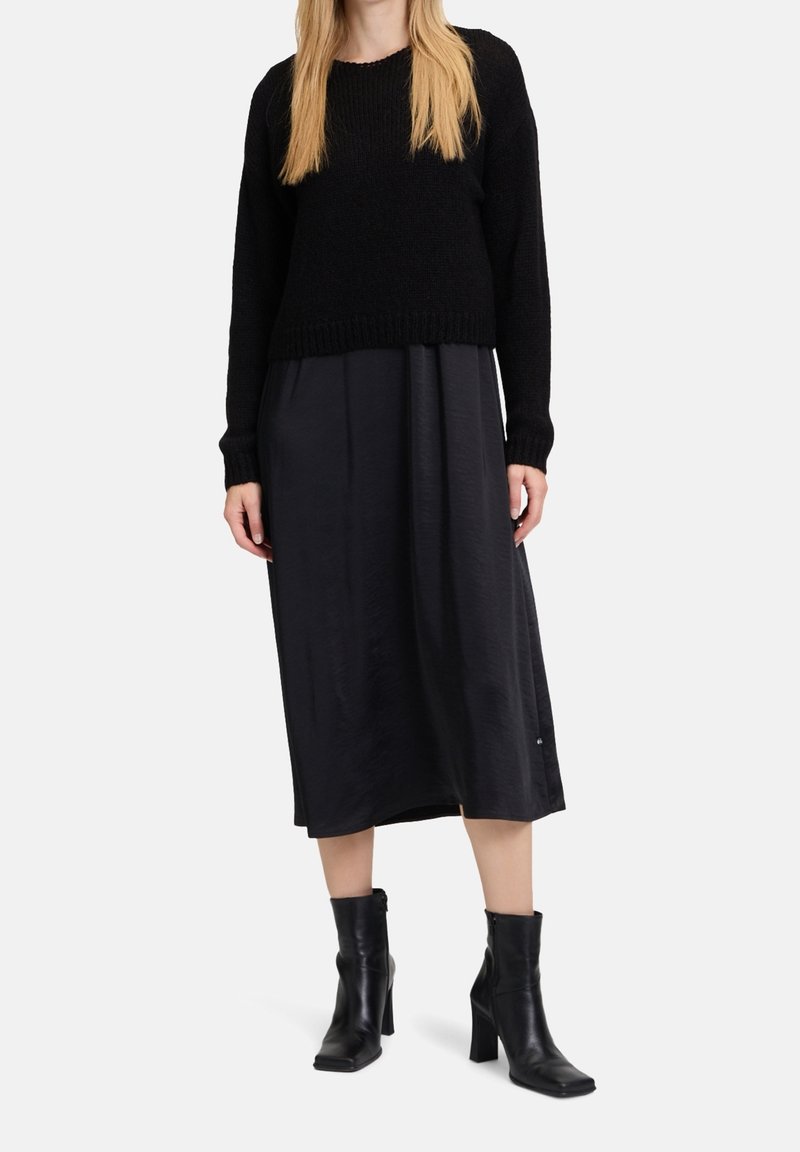 Femme portant un pull en maille noir, une jupe midi noire et des bottines noires à talons, debout devant un fond uni.