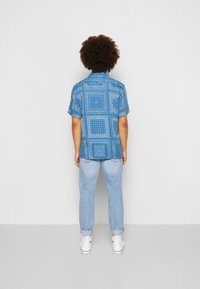 Camicetta blu a maniche corte con un design a motivo, abbinata a jeans azzurri chiari e sneakers bianche. La camicia presenta varie forme geometriche.