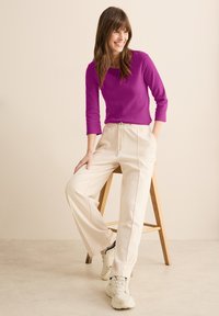 Lila tight topp med trekvartslånga ärmar, som matchas med krämfärgade joggerbyxor med elastiskt midjeband och sidofickor. Sneakers fulländar looken.