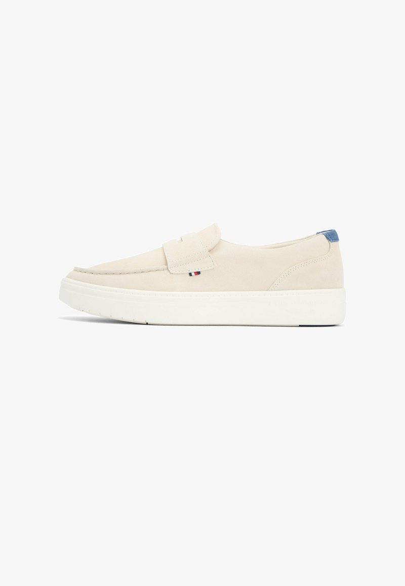 Mocasín beige sin cordones con suela blanca gruesa y pequeño detalle de tela en rojo, blanco y azul cerca de la costura, mostrado de perfil sobre fondo blanco.