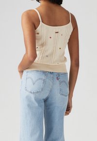 Top de punto beige sin mangas con patrón de trenzas, decorado con bordados de estrellas multicolores; combinado con jeans de tiro alto en azul claro.