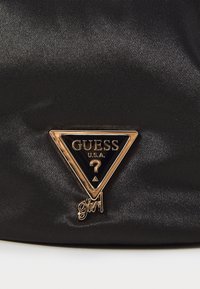 Schwarze Satin-Ledertasche mit einem goldenen Dreieckslogo, auf dem "GUESS" und "girl" am unteren Rand stehen, das mit einem glänzenden Finish hervorgehoben wird.