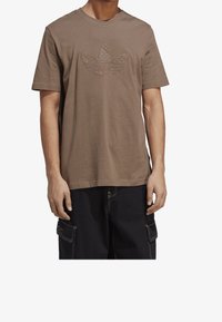 Muž v hnědém tričku s krátkým rukávem s embosovaným logem Adidas trefoil, doplněném černými cargo kalhotami s bílým prošíváním.