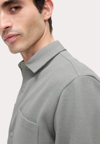 Camisa gris texturizada con cuello, cierre de botones y un bolsillo en el pecho. La tela parece tejida con un patrón sutil, destacando su diseño estructurado.