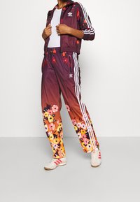 Floral tepláková souprava Adidas v burgundy a oranžové barvě s bílými pruhy. Bunda má zip a stojáček; kalhoty mají květinový vzor a kapsy.